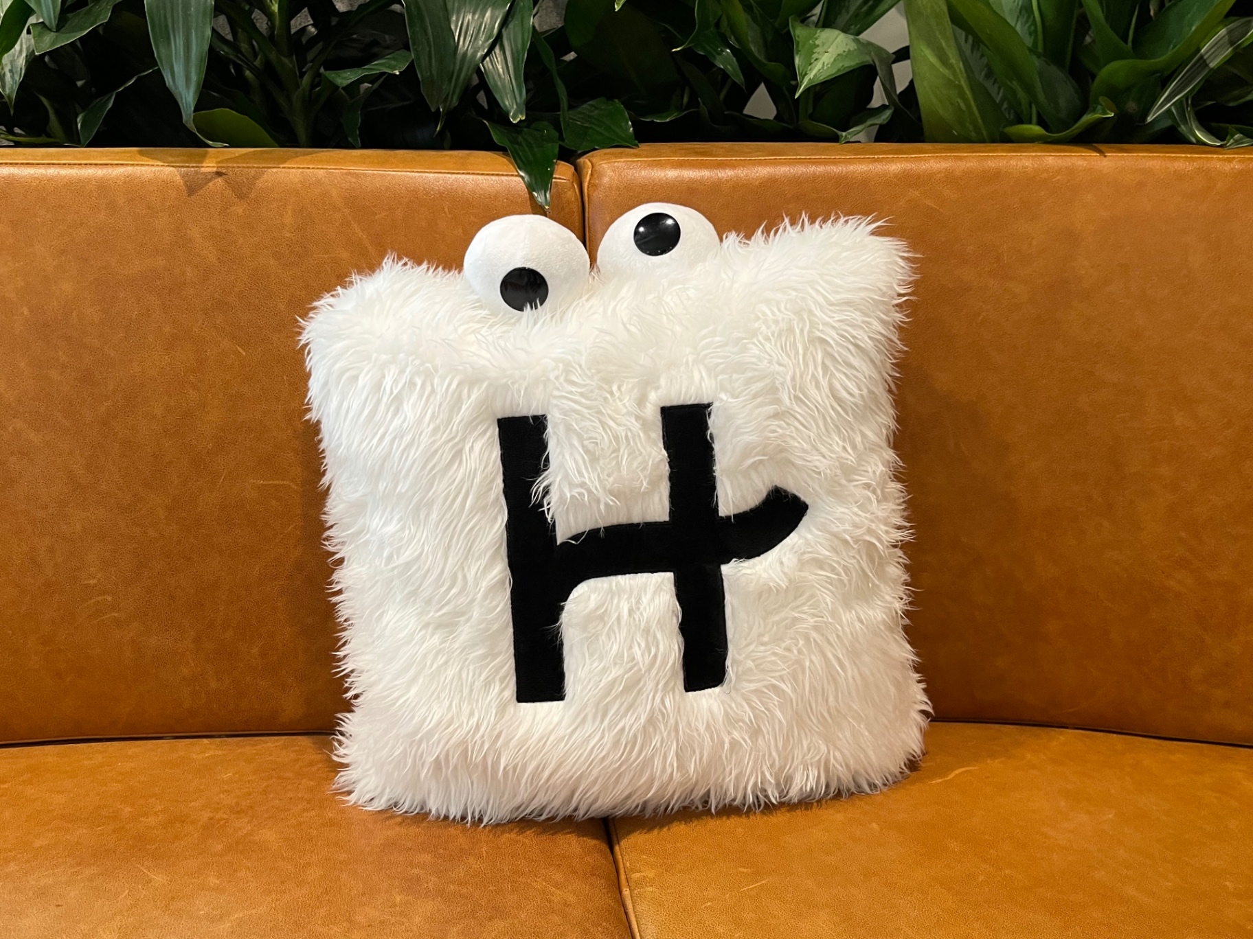 A fuzzy white Hinge H pillow with googly eyes sitting on a leather couch